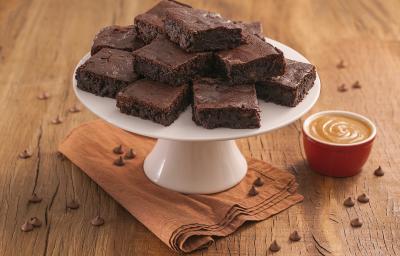 Fotografia em tons de marrom e vermelho de uma bancada de madeira com um paninho, sobre ele um prato branco com pedaços de brownie. Ao lado um potinho com Moça de colher doce de leite.