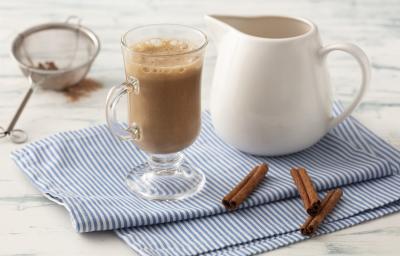 Fotografia em tons de azul em uma bancada de madeira clara com um pano azul com listras finas e ao centro uma taça com o cappuccino e ao lado uma jarrinha branca. Ao lado, três paus de canela e um difusor com o chocolate em pó.