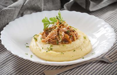 Fotografia em tons de cinza, branco e amarelo, prato branco ao centro com polenta, frango desfiado e salsa, ao lado um garfo base, sobre toalha com listras brancas e cinza e ao fundo guardanapo cinza.