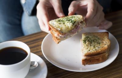 Fotografia de um sanduíche aberto com uma moça segurando a metade do lanche, que tem presunto, lombinho defumado, molho e queijo gruyère gratinado por cima. O sanduíche está em um prato raso branco de vidro ao lado de uma xícara branca de café cheia.