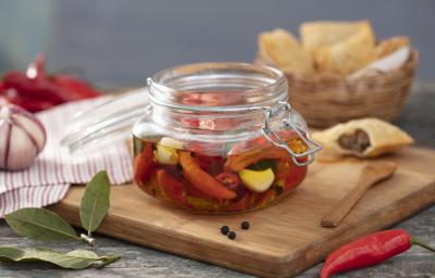 Fotografia em tons de vermelho em uma bancada de madeira clara, um pano branco com listras vermelhas, uma tábua de madeira com um pote hermético de vidro em cima e com as pimentas em conserva dentro dele. Ao fundo, uma cesta com pasteis e pimentas.