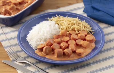 Fotografia em tons de azul em uma bancada de madeira com um jogo americano branco com listras azuis, um prato fundo azul com o strogonoff de salsicha, arroz branco e batata palha. Ao fundo, um recipiente com mais strogonoff de salsicha.