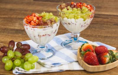 Fotografia em tons de azul e bege em uma bancada e madeira escura, um pano listrado em azul e bege, duas taças de vidro com a sobremesa de creme com morangos e uvas picados. Ao lado, um potinho com morangos em cima e cachos de uvas.