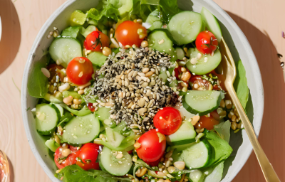 Fotografia mostra uma salada verde com tomate, pepino, grãos, sementes e um pouco de gergelim preto espalhado pela salada