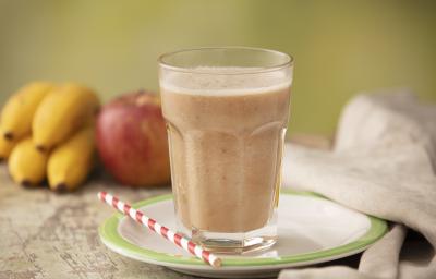 Fotografia em tons de cinza de uma bancada cinza com um prato branco com borda verde sobre ele um canudo listrado branco e vermelho e um copo de vidro com a vitamina de banana e maçã. Ao fundo um cacho de banana, uma maçã e um paninho cinza.