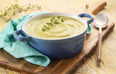 Foto da receita de creme de brócolis em uma panela azul em cima de uma tábua de madeira com um pano azul claro apoiado na panela. Em cima da tábua ainda há uma colher longa prateada e esses elementos estão dispostos em cima de uma mesa de madeira.