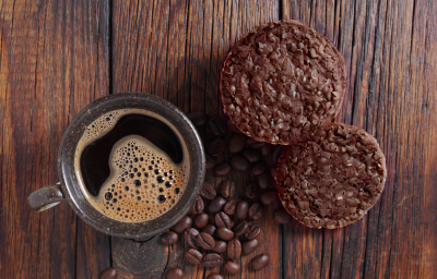 Foto vista de cima da receita de cookies de aveia e café, servidos em uma bancada de madeira, ao lado de uma xícara de café preparada com alguns grãos de café espalhados