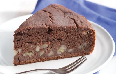 Fotografia em tons de azul em uma mesa de madeira branca com um pano azul, um prato branco raso com uma fatia de bolo de chocolate com banana.