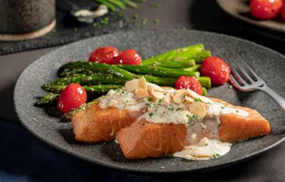 Foto de um prato escuro com a receita de salmão grelhado, acompanhado de aspargos e tomates cereja. Ao fundo há outro prato aparecendo, alguns aspargos um copo preto pequeno com molho dentro