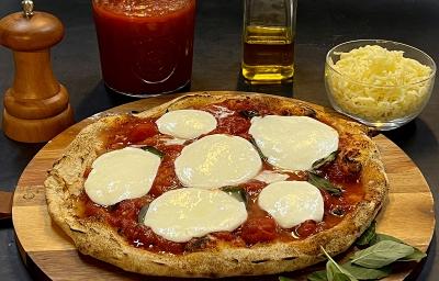 Foto da receita de pizza napolitana servida sobre uma tábua de madeira redonda com folhas manjericão ao lado. Ao fundo, há um bowl de vidro com muçarela, um vidro de azeite, um moedor de sal e um vidro de molho pomodoro