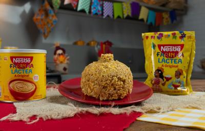Fotografia de um prato pequeno, raso e vermelho apoiado em alguns panos de cor bege, um vermelho e outro amarelo quadriculado com branco. No prato tem uma receita de cuscuz com farinha láctea, ao lado do prato tem as embalagens da marca. Fotografia de um prato pequeno, raso e vermelho apoiado em alguns panos de cor bege, um vermelho e outro amarelo quadriculado com branco. No prato tem uma receita de cuscuz com farinha láctea, ao lado do prato tem as embalagens da marca.