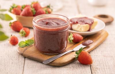 Foto em tons de vermelho da receita de geleia cremosa de morango servida em um recipiente de vidro com uma faca arredonda prateada ao lado e morangos decorativos. Ao fundo há um prato com um pão e a geleia por cima, além de uma cesta com morangos