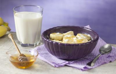 Fotografia em tons de roxo em uma bancada de madeira com um pano roxo com bolinhas brancas, um pote grande e fundo roxo com as bananas cobertas com aveia e meia. Ao lado, um copo de leite, um potinho com o mel.