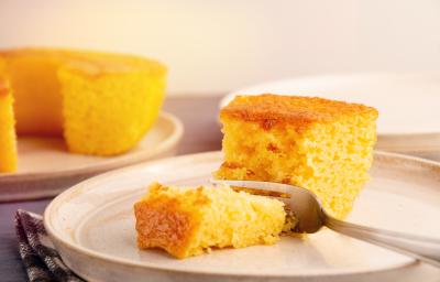 Fotografia de um prato grande de vidro com uma fatia grande de bolo de milho e um garfo cortando um pedaço. No fundo tem o bolo inteiro, sem essa fatia, sobre um apoio raso de cor clara.