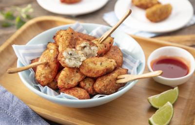 Fotografia em tons de bege em uma bancada de madeira clara, uma tábua de madeira, um pano azul, um prato redondo fundo com vários bolinhos de peixe, um potinho branco pequeno com molho de pimenta e pedaços de limão. Ao fundo, pratinhos brancos com bolinho