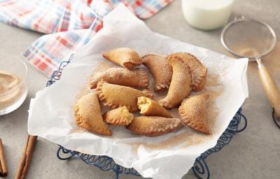 Foto da Receita de Pastel de Grão-de-bico. Observa-se um prato com papel absorvente e os pasteizinhos em cima polvilhados de canela e açúcar. Ao lado esquerdo, dois paus de canela e, ao direito, uma peneira com.