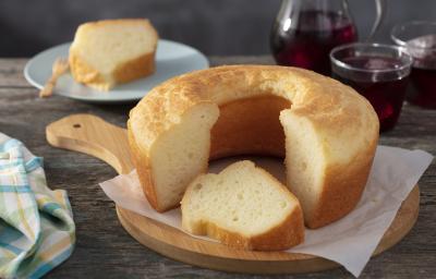 Fotografia em tons de amarelo em uma bancada de madeira escura, um pano azul com listras amarelas, uma tábua de madeira redonda com o bolo de pão de queijo em cima dele. Ao fundo, um prato pequeno com um pedaço do bolo em cima dele.