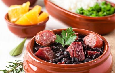 Fotografia de um recipiente médio marrom clássico com uma feijoada com caldo de bacon, linguiça e bacon com uma folha de salsinha. Ao fundo tem outros recipientes iguais, porém menores, com laranja, arroz, couve e farofa.