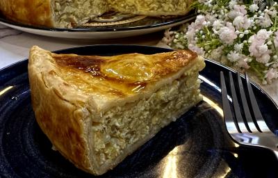 Foto da receita de Torta de Cebola e Ovos. Observa-se um pedaço grande de torta em um prato preto com decoração de flores brancas.