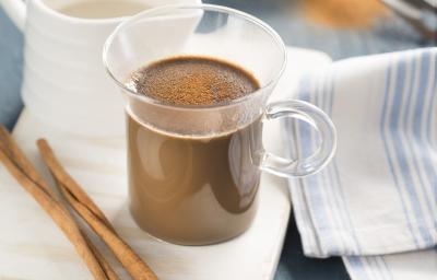 fotografia em tons de branco, marrom e azul tirada de uma xicara transparente com o Cappuccino dentro, e ao lado direito contém uma xicara branca e à frente canela.