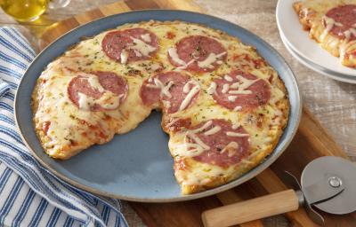 Foto da receita de pizza caseira com massa de batata com rodelas de salame por cima sobre uma base circular de cerâmica com um cortador de pizza prateado ao lado sobre uma tábua de madeira. Ao lado há um paninho azul listrado e um vidro de azeite