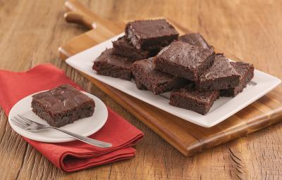 Fotografia em tons de marrom e vermelho de uma bancada de madeira com uma tábua de madeira, sobre ele um papel manteiga com pedaços de brownie. Ao  fundo um prato com um pedaço de brownie e um garfo.