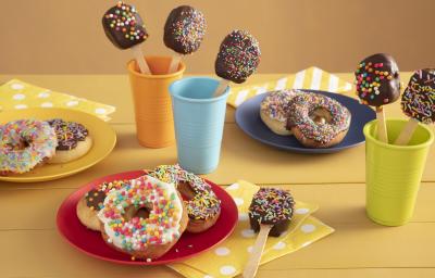 Fotografia em tons de amarelo em uma bancada de madeira amarela com três copos coloridos, azul, laranja e verde com pirulitos de chocolate dentro dele. Ao centro, um prato vermelho com três donuts cobertos com granulados coloridos.