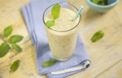 Fotografia em tons de marrom e azul de uma bancada de madeira com um paninho azul, sobre ele um copo com smoothie com um canudo. Ao lado folhinhas de hortelã e um recipiente redondo com hortelã.