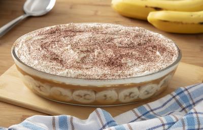 Foto da receita de pavê de banoffee servida em uma travessa de vidro grande sobre uma tábua de madeira em cima de uma mesa de madeira com um paninho quadriculado ao lado