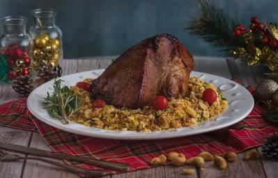 Foto da receita de pernil com farofa crocante, servido em um prato branco redondo, sobre uma mesa de madeira toda decorada com itens natalinos. Há algumas castanhas de caju espalhadas na mesa e um conjunto de talheres à frente do prato