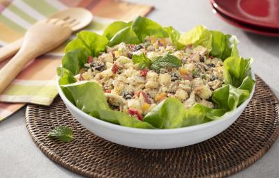 Fotografia em tons de verde em uma bancada de madeira, um pano com listras coloridas, um jogo americano redondo de vime escuro e um recipiente branco redondo fundo com a salada marroquina com vários legumes dentro dela.