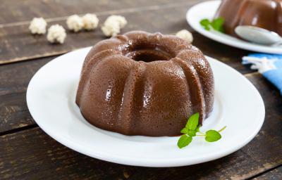 Fotografia de um pudim pequeno de chocolate meio amargo e folhas de hortelã que estão em cima de um prato raso de vidro na cor branco. Ao fundo, o mesmo pudim sobre um prato, um pano azul e algumas pipocas sobre uma mesa de madeira de tom escuro.