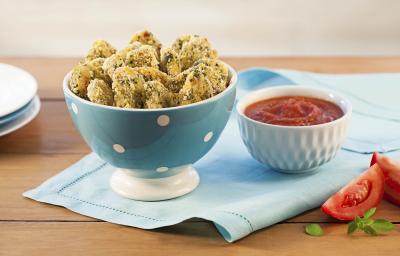 Fotografia em tons de marrom e azul claro de uma mesa de madeira vista de frente, contém um pano cor azul claro e por cima um recipiente redondo cor azul com bolinhas brancas com pedaços de nuggets, ao lado um pote branco com molho de tomate.