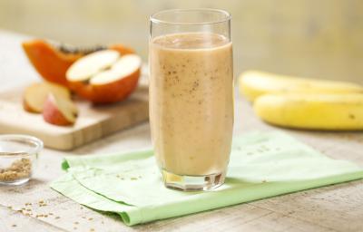 Fotografia em tons de rosa, verde e branco, com copo ao centro com vitamina sobre guardanapo verde, no entorno duas bananas, maçã cortada em pedaços e um pedaço de mamão sobre peça de madeira, potinho com linhaça, tudo sobre bancada em tons de branco.