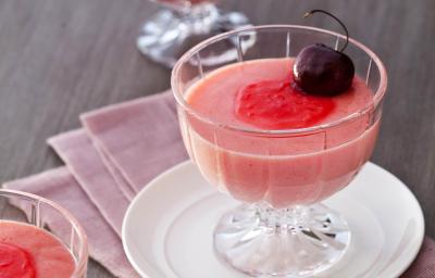 Fotografia em tons de vermelho em uma mesa de madeira com um pano rosa escuro e um prato pequeno branco apoiado nele. Em cima do prato, uma taça de vidro com a mousse de cereja e uma cereja por cima para enfeita. Ao lado e ao fundo, mais duas taças.