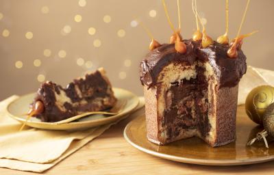 Fotografia em tons de amarelo em uma bancada de madeira clara, um pano amarelo e decoração natalina de fundo. Ao centro, um prato dourado com o panettone de avelã recheado sem um pedaço. Ao lado, um prato com a fatia do panettone.