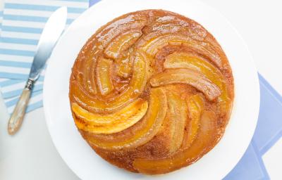 Fotografia em tons de azul e branco de uma bancada branca com um paninho azul, sobre ela um prato redondo branco com um bolo de banana. Ao lado um paninho listrado e uma faca.