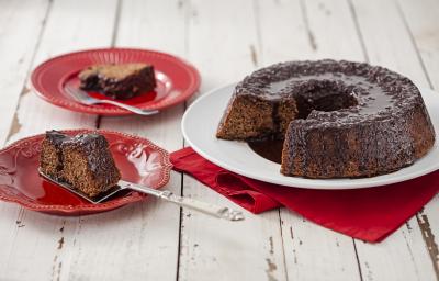 Fotografia em tons de vermelho em uma bancada de madeira clara com um prato branco ao centro com o bolo de chocolate cortado ao meio e um paninho vermelho embaixo. Ao lado, dois pratos vermelhos com uma fatia de bolo em cada um.