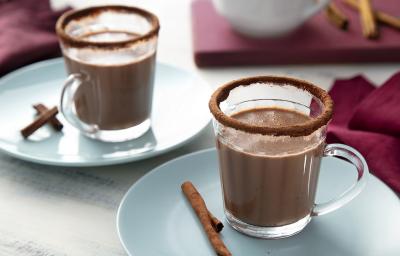 Fotografia em tons de marrom, em uma bancada de madeira de cor branca. Ao centro, um pires azul claro contendo uma xícara de chocolate quente com uma canela em pau ao lado. Ao fundo, um pano roxo e um pires com outra xícara.