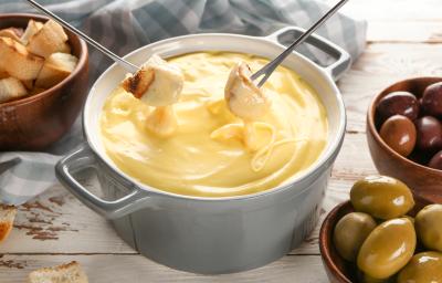 Fotografia em tons de branco com uma panela de fondue ao centro. Dentro da panela existe um fondue de queijo. Dois pedaços de pão estão sendo mergulhados no fondue de queijo. Ao fundo existe recipientes com azeitona e pedaços de pão.