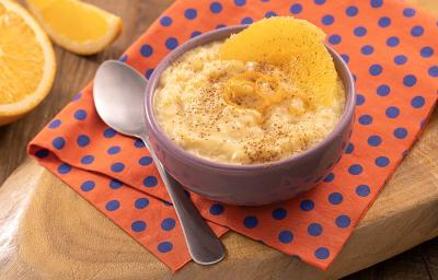 Foto em tons de laranja da receita de arroz-doce de laranja servida em um bowl de porcelana lilás sobre um pano laranja com bolinhas azuis com uma colher prateada ao lado. Ao fundo há laranjas cortadas sobre uma tábua de madeira