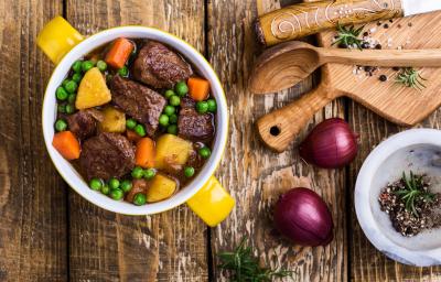 Fotografia vista de cima de um cozido de carne com ervilhas frescas, legumes e caldo de carne. Ao lado, duas cebolas roxas, uma tábua e colher de madeira e alguns temperos que estão dentro de um refratário fundo e pequeno, sobre uma mesa de madeira.