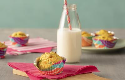 Fotografia em tons de rosa, em uma bancada de madeira de cor cinza. Ao centro, uma tábua de madeira com um pano rosa em cima. Em cima do pano está o muffin e ao lado há um pote de vidro contendo leite. AO fundo alguns muffins espalhados.