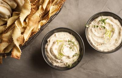 fotografia em tons de cinza, branco e pardo de uma bancada cinza vista de cima. Contém dois potinhos redondos e pretos com uma pasta de creme, e ao lado biscoitinhos para servir junto ao creme.