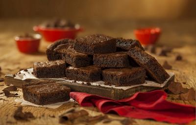 Fotografia em tons de marrom em uma bancada de madeira de cor marrom. Ao centro um pano vermelho contendo uma tábua de madeira com os brownies em cima. Ao redor, há vários pedaços de chocolate espalhados.