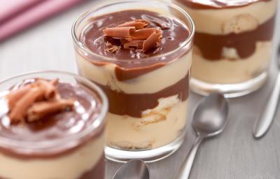 Fotografia em tons de marrom em uma bancada de madeira clara com um pano rosa de fundo e três taças de vidro com pavê de chocolate com maracujá e raspas de chocolate para enfeitar.