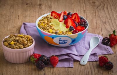 Fotografia em tons de lilás em uma bancada de madeira clara, um bowl colorido com o iogurte de abacate com frutas vermelhas e granola decorando. Ao lado, um potinho com granola e frutas espalhadas na bancada.