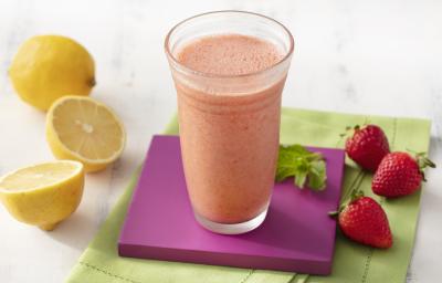 Fotografia em tons de branco, roxo e verde de uma bancada de madeira branca e um paninho verde, sore ele um apoio de copo quadrado roxo com um copo de vidro com o suco de morango. Ao lado três morangos e dois limões.