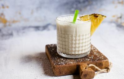 Fotografia em tons claros de uma bebida de neston com suco de abacaxi que está em um copo pequeno de vidro. Na borda do copo tem abacaxi e um canudo verde.