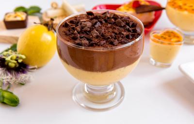 Fotografia de uma sobremesa de maracujá, chocolate e biscoito em uma taça, que está sobre uma mesa branca. Ao fundo, diversas opções de mousse de maracujá, a fruta inteira e uma tábua de madeira clara.